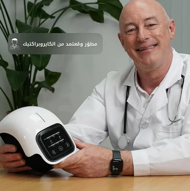 Man in a white coat holding a white device with a screen, sitting at a desk with a plant in the background.